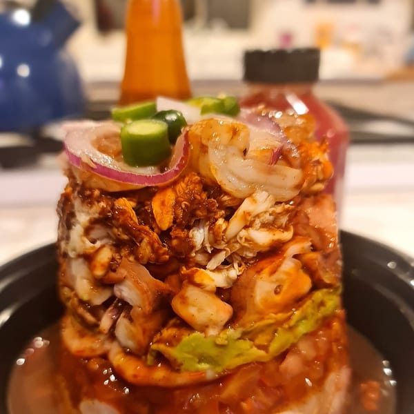 A tall, layered seafood cocktail with shrimp, avocado, red onion, and jalapeños in a dark bowl.