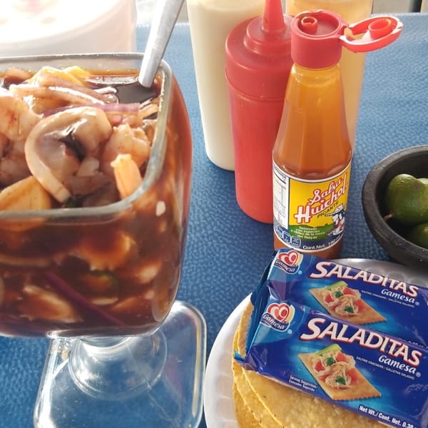 A glass of shrimp and mushroom ceviche with hot sauce, limes, and crackers on a blue table.