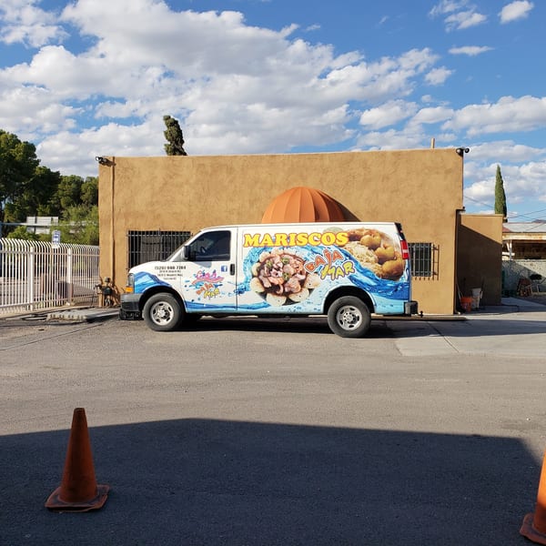 A white food truck with "Mariscos" and seafood graphics is parked in front of a tan building.