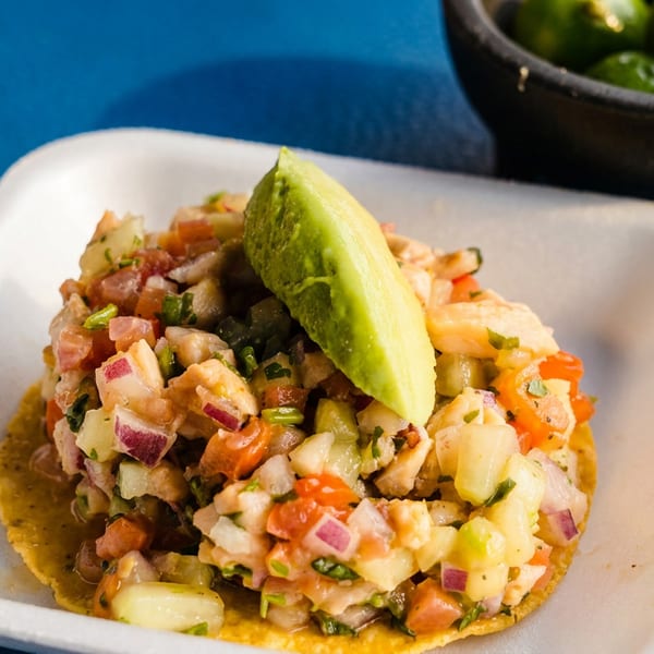 A vibrant ceviche tostada with avocado, tomatoes, and onions on a white plate, with limes in the background.