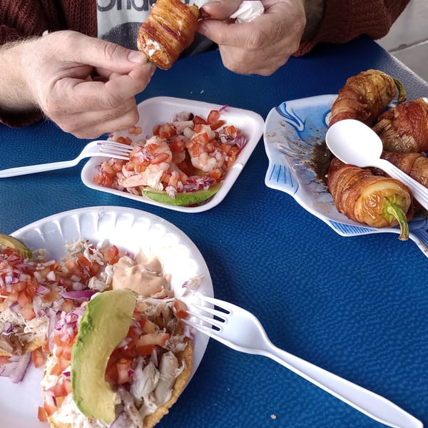 Hands holding a bacon-wrapped jalapeño, with shrimp ceviche and tostadas on a blue table.