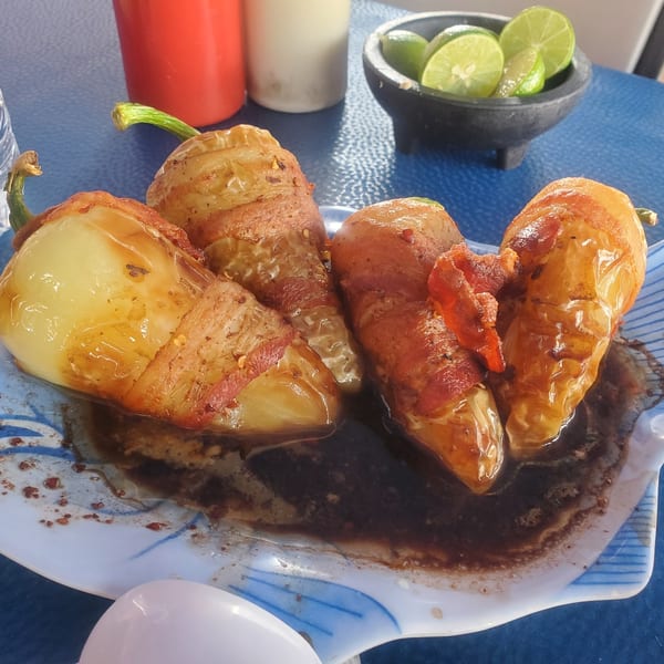 Four bacon-wrapped peppers in a dark sauce on a fish-shaped plate, with limes in the background.